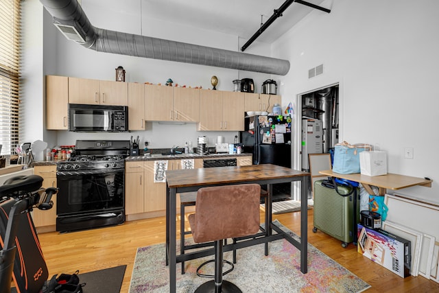 kitchen with black appliances, light brown cabinetry, light hardwood / wood-style flooring, and a towering ceiling