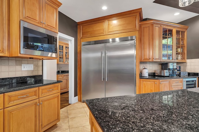 kitchen with built in appliances, wine cooler, light tile patterned flooring, dark stone counters, and glass insert cabinets