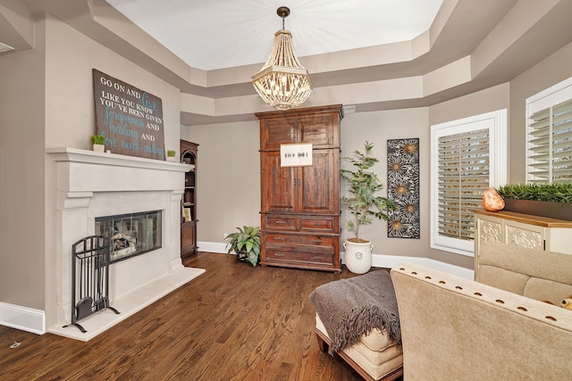 living area featuring baseboards, a raised ceiling, dark wood finished floors, a glass covered fireplace, and a notable chandelier