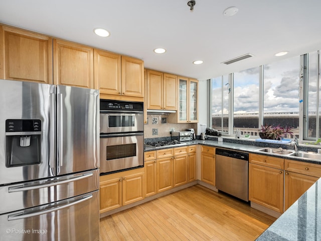 kitchen with light wood-type flooring, appliances with stainless steel finishes, light brown cabinetry, sink, and dark stone countertops