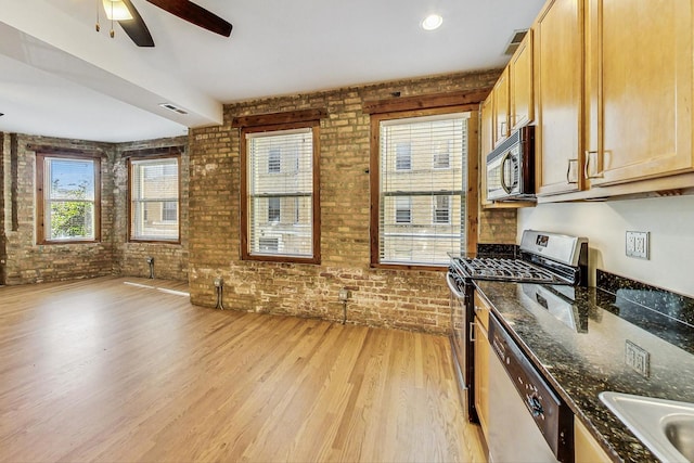 kitchen with stainless steel appliances, dark stone counters, ceiling fan, light hardwood / wood-style flooring, and brick wall