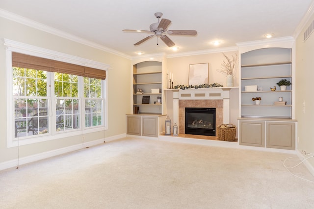 unfurnished living room featuring a tiled fireplace, built in shelves, ornamental molding, ceiling fan, and light colored carpet