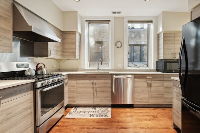 kitchen featuring light hardwood / wood-style floors, wall chimney exhaust hood, black appliances, sink, and light brown cabinets