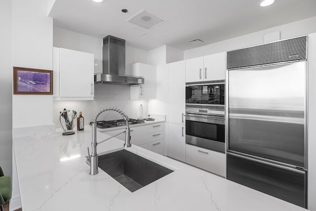 kitchen with stainless steel appliances, sink, light stone countertops, white cabinets, and wall chimney range hood