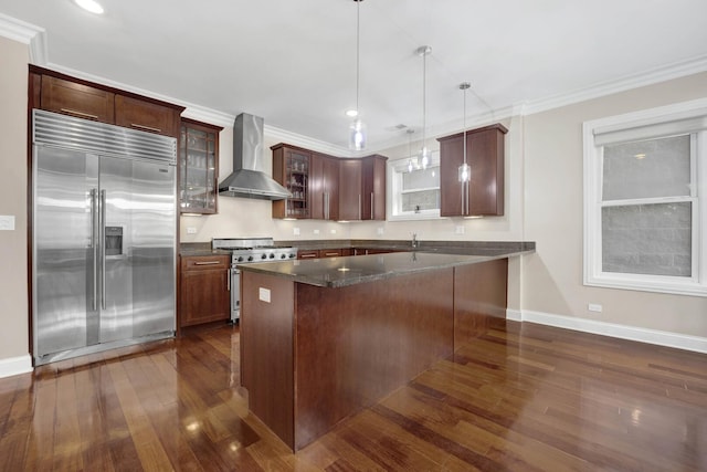 kitchen with wall chimney exhaust hood, high quality appliances, glass insert cabinets, ornamental molding, and dark wood-style flooring