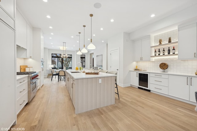 kitchen with wine cooler, stainless steel range, an island with sink, white cabinets, and decorative light fixtures