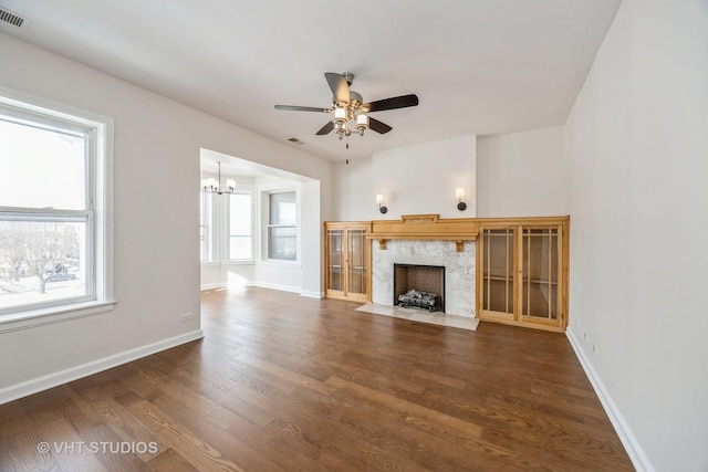 unfurnished living room featuring ceiling fan with notable chandelier, a premium fireplace, visible vents, baseboards, and dark wood finished floors