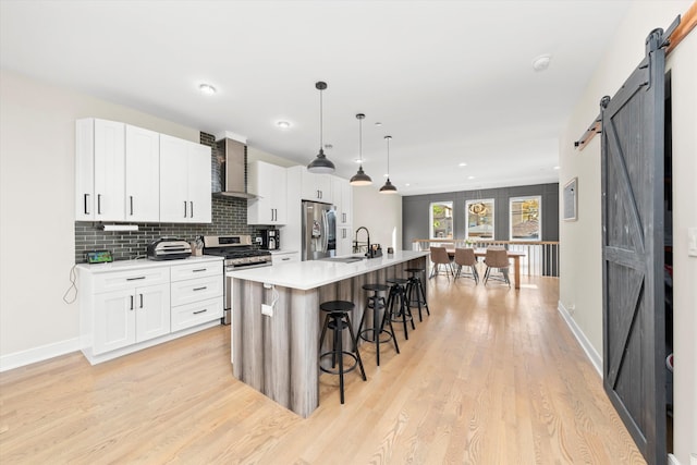 kitchen featuring stainless steel appliances, wall chimney range hood, a barn door, hanging light fixtures, and a kitchen island with sink