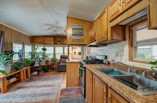 kitchen with ceiling fan, sink, stainless steel gas stove, lofted ceiling, and wood walls