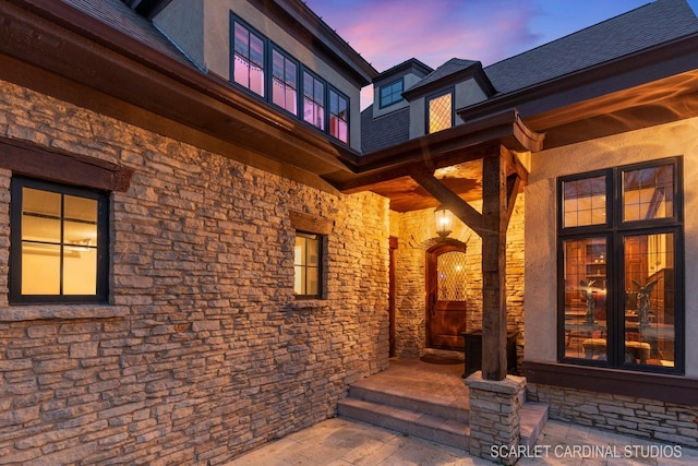 exterior entry at dusk featuring stone siding, stucco siding, and roof with shingles