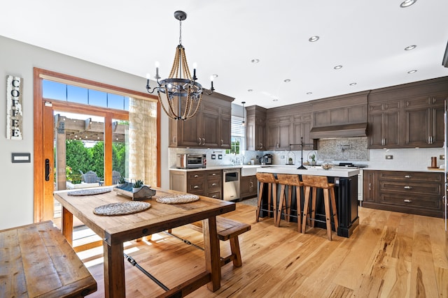 kitchen with backsplash, dark brown cabinets, exhaust hood, an inviting chandelier, and light hardwood / wood-style flooring