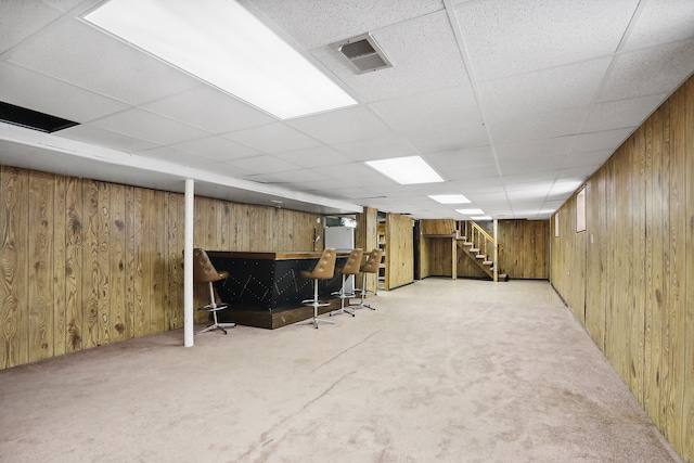 basement with light colored carpet, a paneled ceiling, wooden walls, and indoor bar