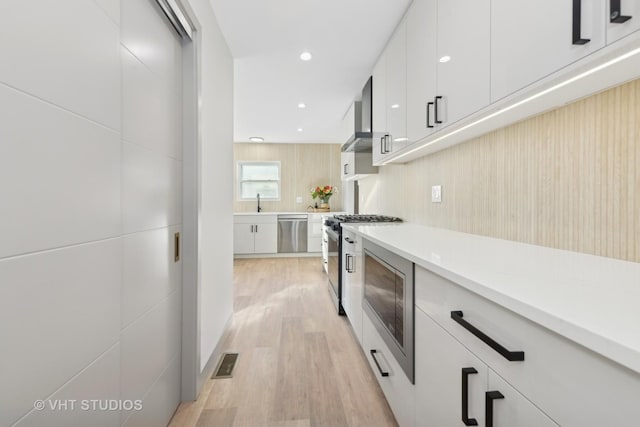 kitchen with sink, appliances with stainless steel finishes, white cabinetry, wall chimney exhaust hood, and light wood-type flooring