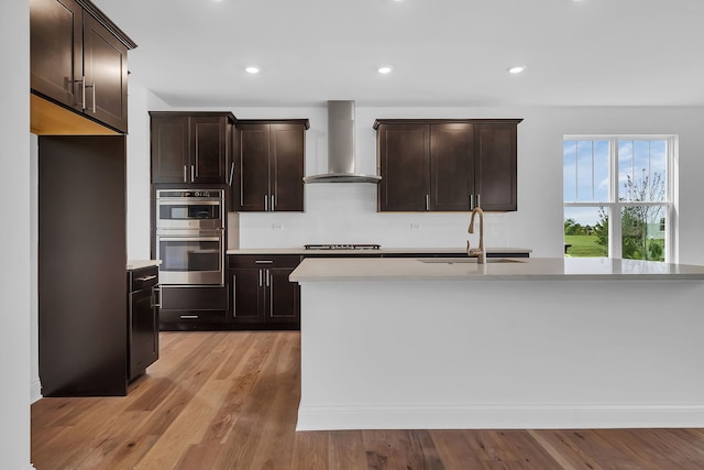 kitchen with sink, an island with sink, stainless steel double oven, wall chimney range hood, and light wood-type flooring