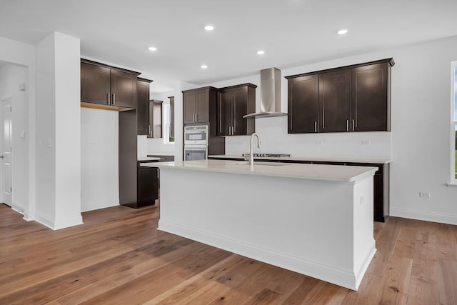 kitchen featuring a center island with sink, wall chimney exhaust hood, sink, light hardwood / wood-style floors, and double oven