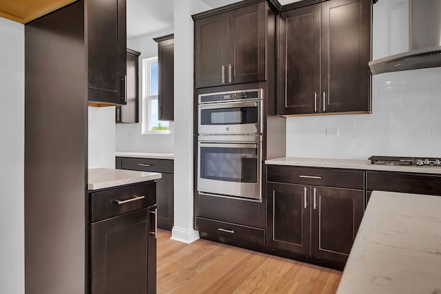 kitchen with appliances with stainless steel finishes, dark brown cabinetry, decorative backsplash, wall chimney exhaust hood, and light hardwood / wood-style flooring