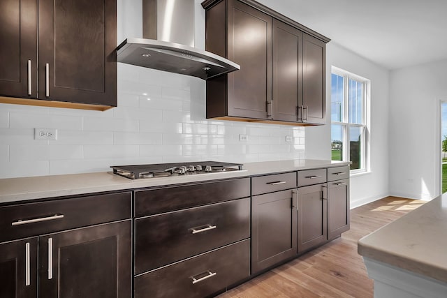 kitchen with stainless steel gas cooktop, wall chimney exhaust hood, dark brown cabinets, light hardwood / wood-style flooring, and decorative backsplash