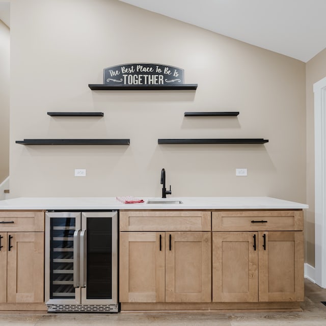 bar featuring sink, beverage cooler, vaulted ceiling, light brown cabinetry, and light wood-type flooring