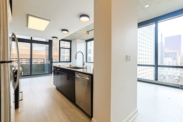 kitchen with stainless steel appliances, sink, a wall of windows, dark stone countertops, and light hardwood / wood-style flooring