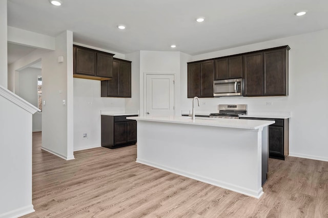 kitchen featuring sink, an island with sink, light hardwood / wood-style floors, dark brown cabinets, and appliances with stainless steel finishes