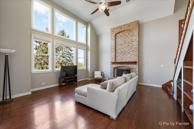 living room featuring dark wood-style flooring, a ceiling fan, a brick fireplace, high vaulted ceiling, and baseboards