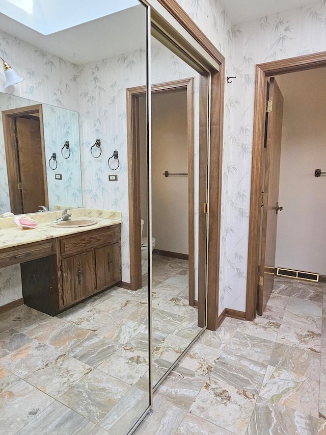 bathroom featuring a skylight, wallpapered walls, baseboards, toilet, and vanity