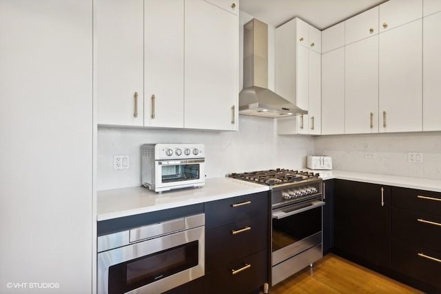 kitchen featuring appliances with stainless steel finishes, tasteful backsplash, wall chimney exhaust hood, light hardwood / wood-style flooring, and white cabinets