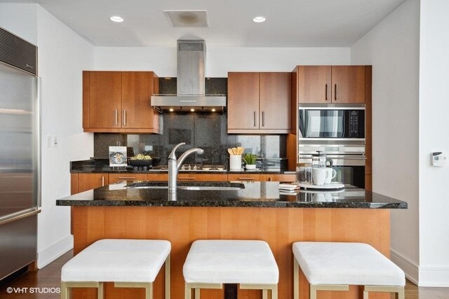 kitchen with built in appliances, wall chimney exhaust hood, dark wood-type flooring, and a breakfast bar