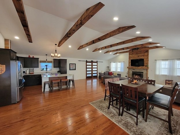 dining room featuring a barn door, a stone fireplace, lofted ceiling with beams, a chandelier, and light wood-type flooring