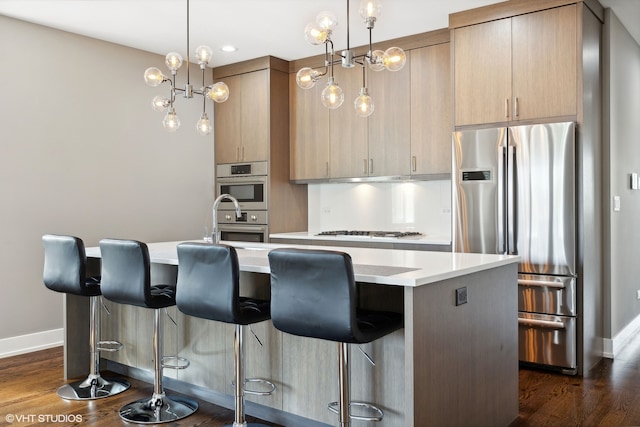 kitchen with a center island with sink, hanging light fixtures, stainless steel appliances, and light brown cabinets