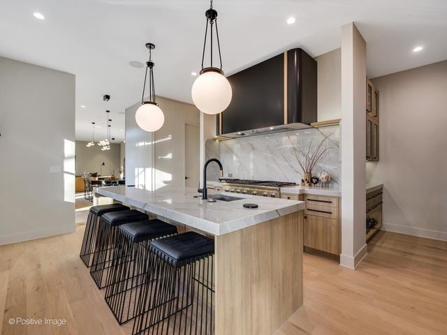 kitchen featuring sink, wall chimney exhaust hood, pendant lighting, a kitchen island with sink, and light wood-type flooring