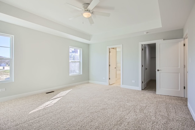 unfurnished bedroom featuring ceiling fan, a spacious closet, connected bathroom, a tray ceiling, and light colored carpet