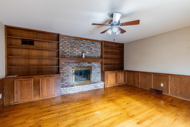 unfurnished living room with wood walls, a brick fireplace, light hardwood / wood-style flooring, built in shelves, and ceiling fan