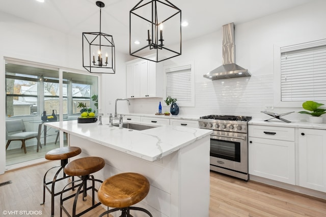 kitchen with white cabinets, stainless steel stove, wall chimney exhaust hood, an island with sink, and decorative light fixtures