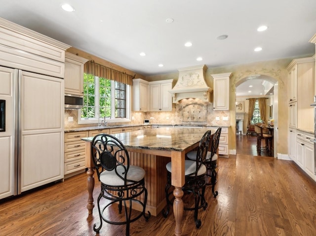 kitchen featuring arched walkways, light stone counters, a kitchen breakfast bar, dark wood-type flooring, and premium range hood