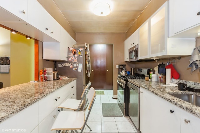 kitchen with light tile patterned floors, light stone counters, white cabinets, appliances with stainless steel finishes, and glass insert cabinets