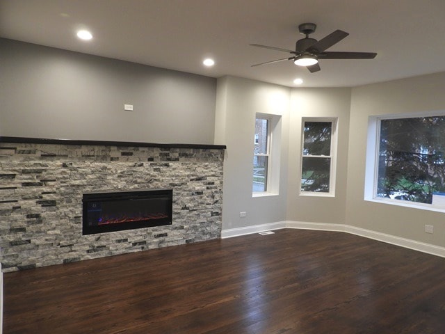 unfurnished living room with ceiling fan, a healthy amount of sunlight, dark hardwood / wood-style flooring, and a stone fireplace