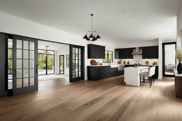 kitchen with french doors, wall chimney exhaust hood, light hardwood / wood-style floors, a kitchen island with sink, and a breakfast bar