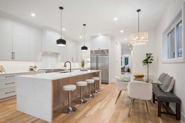 kitchen featuring light wood-type flooring, a kitchen island with sink, decorative light fixtures, stainless steel built in fridge, and white cabinets