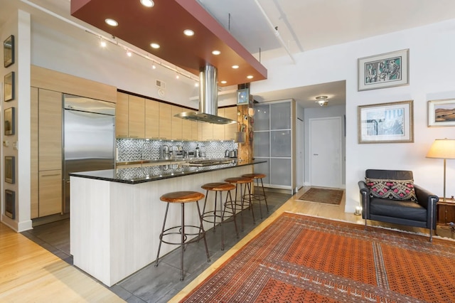 kitchen featuring a kitchen breakfast bar, backsplash, island exhaust hood, stainless steel built in fridge, and hardwood / wood-style flooring