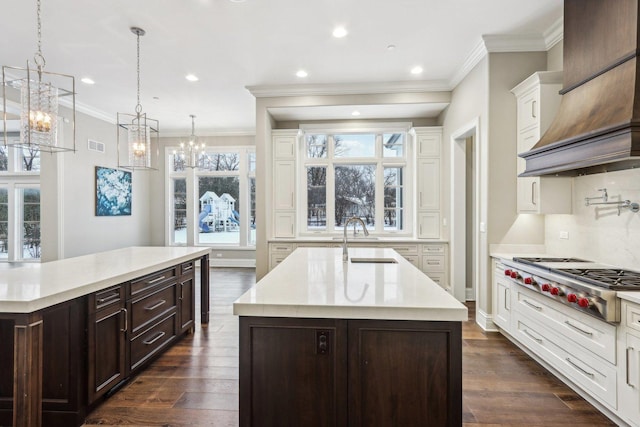 kitchen with custom range hood, decorative light fixtures, sink, stainless steel gas stovetop, and a center island with sink