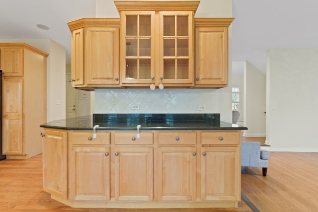 kitchen featuring light wood-style floors, decorative backsplash, light brown cabinetry, dark stone countertops, and glass insert cabinets