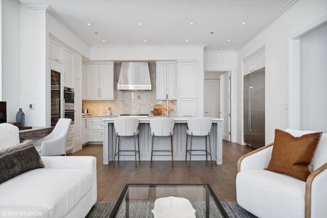 kitchen featuring a breakfast bar, dark wood-type flooring, wall chimney range hood, a center island with sink, and white cabinetry