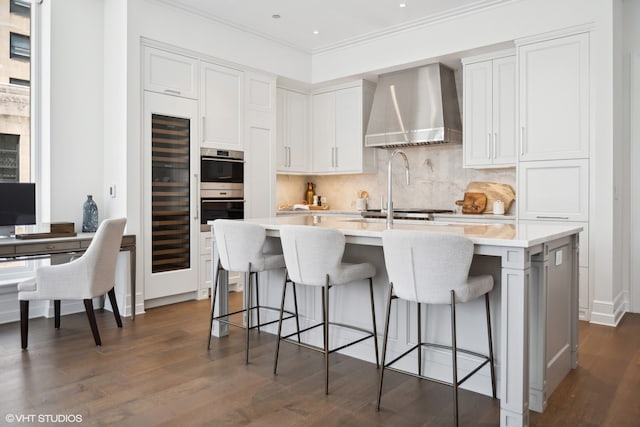 kitchen with dark wood-type flooring, wall chimney range hood, crown molding, an island with sink, and white cabinets