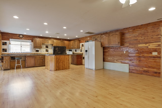 kitchen featuring wooden walls, ceiling fan, light wood-type flooring, white fridge with ice dispenser, and a kitchen island