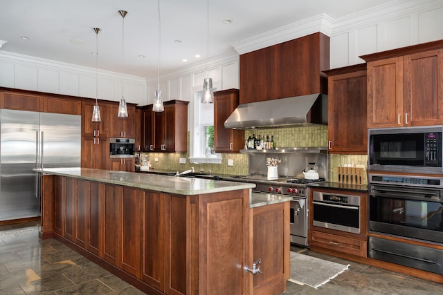 kitchen featuring hanging light fixtures, wall chimney range hood, built in appliances, dark stone counters, and a kitchen island