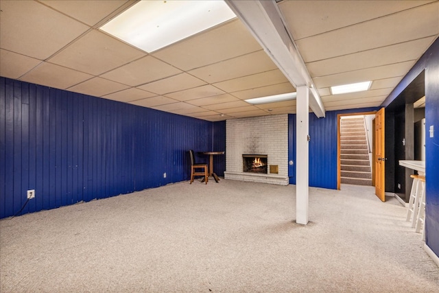 basement featuring carpet, a brick fireplace, a drop ceiling, and wood walls