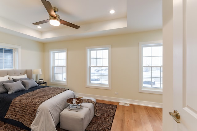 bedroom featuring a tray ceiling, multiple windows, ceiling fan, and light hardwood / wood-style flooring