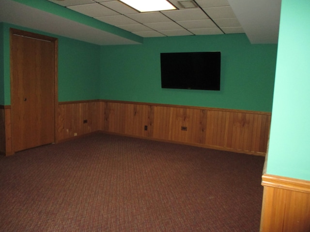 unfurnished room featuring wood walls, a drop ceiling, and dark colored carpet