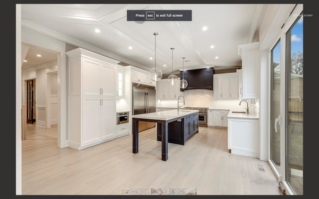 kitchen with high end appliances, white cabinetry, hanging light fixtures, a kitchen island with sink, and ornamental molding